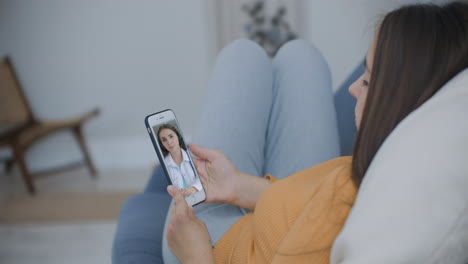 A-woman-lying-on-the-couch-talking-to-a-woman-doctor-on-video-using-a-smartphone.-Remote-medical-consultation-with-a-doctor-via-mobile-phone